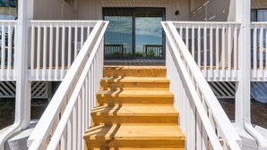 Una escalera de madera que lleva a un porche cubierto con barandas blancas y una entrada con puerta de vidrio.