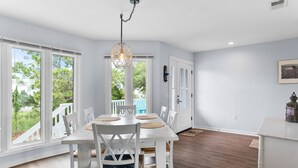 A dining area with a white table and chairs, a hanging light fixture, and large windows with a view of the outdoors. - 3-Bed Harbor Island Home with Ocean Views, Pool Access, and Wildlife Watching! (Saint Helena Island)