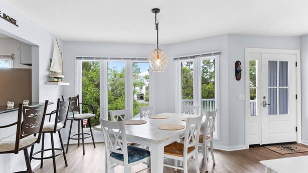 A dining area with a white table and chairs, a hanging light fixture, and large windows offering a view of greenery. - 3-Bed Harbor Island Home with Ocean Views, Pool Access, and Wildlife Watching! (Saint Helena Island)