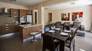 A modern kitchen with a central island, dining area with a table and chairs, and a living area with a sofa and artwork. - Apt. Anna, Modern 2BDR Ayia Napa Apt. with communal pool, Close to the Beach (Ayia Napa)