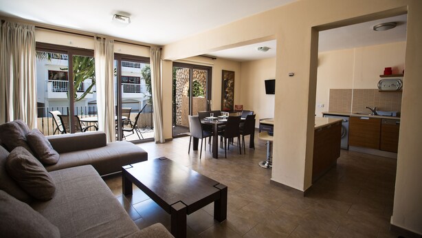 A modern living room with a sofa, a coffee table, and a dining area with chairs and a table. There is a kitchen area with a sink and cabinets. - Apt. Anna, Modern 2BDR Ayia Napa Apt. with communal pool, Close to the Beach (Ayia Napa)