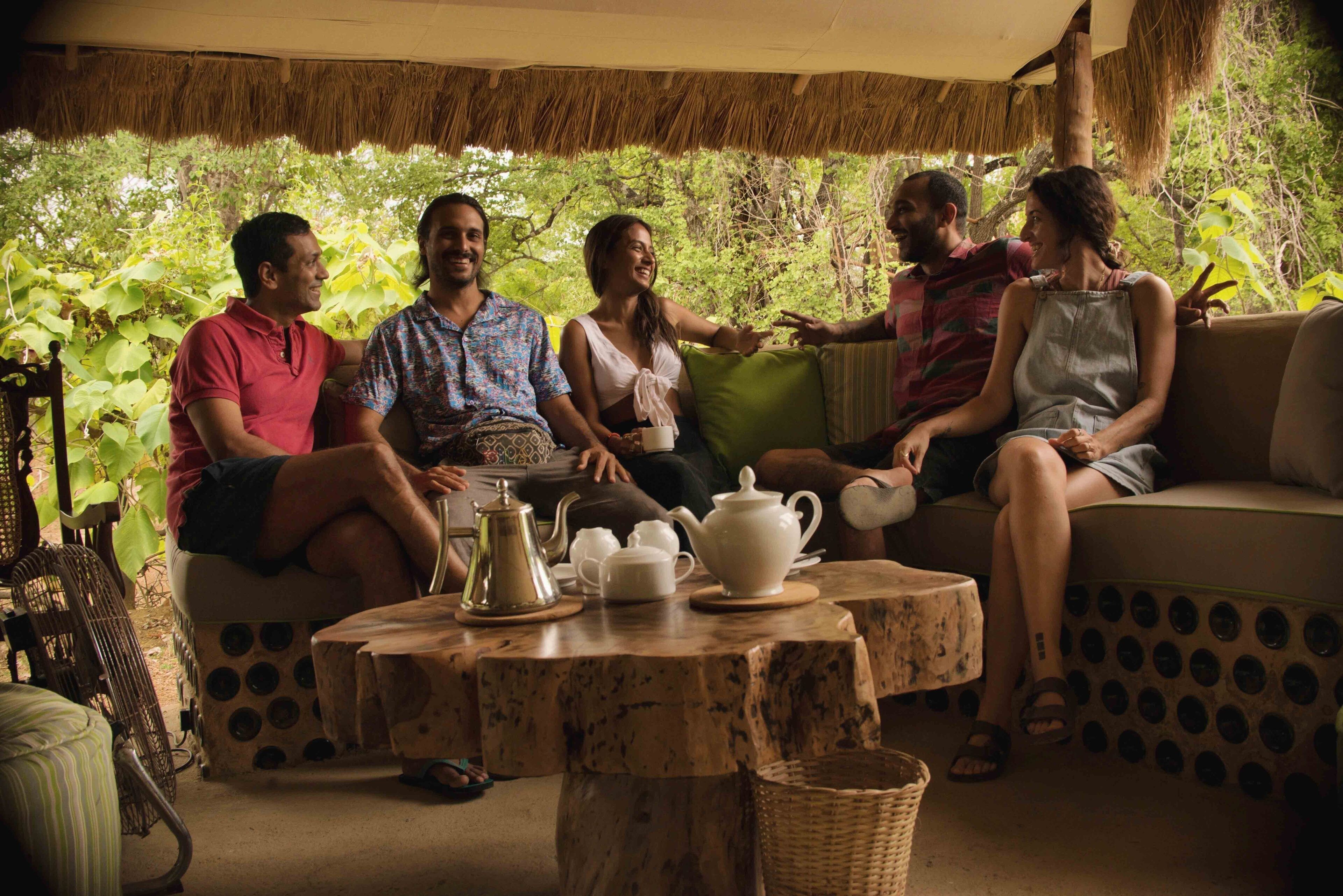 A group of people sitting on a sofa and chairs in a covered outdoor area with a table in front of them.