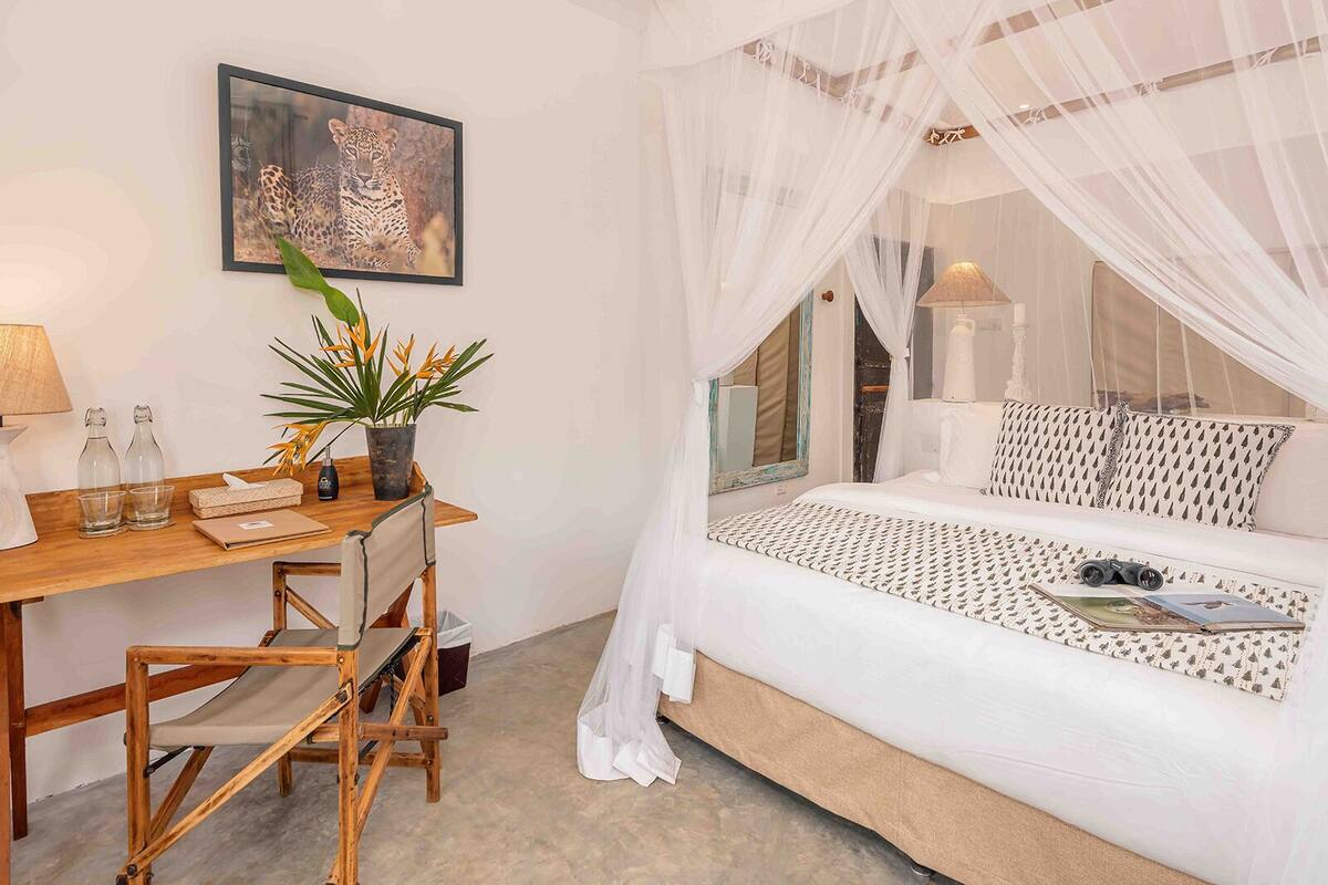 A bedroom with a canopy bed, a desk with a chair, a potted plant, and a framed picture on the wall.