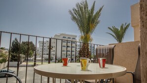 A balcony with a round table and two chairs, overlooking a street with buildings and palm trees. - Apt. Kyla, Lovely 2BDR Ayia Napa Apt with Communal Pool, Close to Nissi Avenue (Ayia Napa)