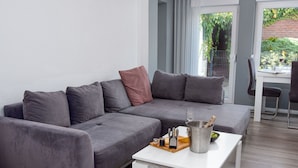 A modern living room with a grey sectional sofa, a white coffee table with a bottle of champagne, and a dining area visible through a glass door. - Lovely apartment for 2 guests with WIFI, TV and terrace (Norden)