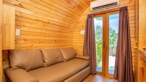 Interior de una cabaña de madera con sofá, unidad de aire acondicionado y puerta corrediza de vidrio con cortinas.