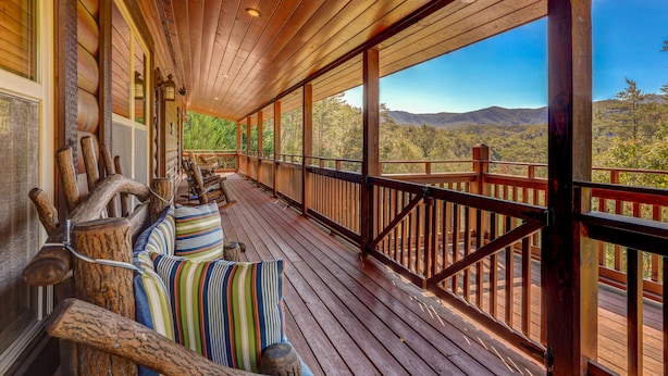 Una terraza de madera con barandilla, un banco con cojines a rayas y vistas a un paisaje montañoso.