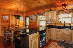 A wooden cabin interior with a kitchen area, wooden cabinets, a dining table with chairs, and a built-in dishwasher. - Gorgeous 5 bedroom cabin with fire pit, hot tub, mountain views, & pool table! (Pigeon Forge)