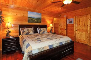 Un dormitorio tipo cabaña de madera con una cama, dos mesitas de noche, un ventilador de techo y una puerta.
