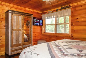A wooden cabin room with a bed, a TV mounted on the wall, and a window with blinds. - Gorgeous 5 bedroom cabin with fire pit, hot tub, mountain views, & pool table! (Pigeon Forge)