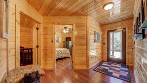 Interior de una cabaña de madera con un pasillo que lleva a un dormitorio, un comedor y una puerta principal con ventanal.