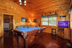 Un interior de madera con mesa de billar, televisor y ventana con vista a árboles.