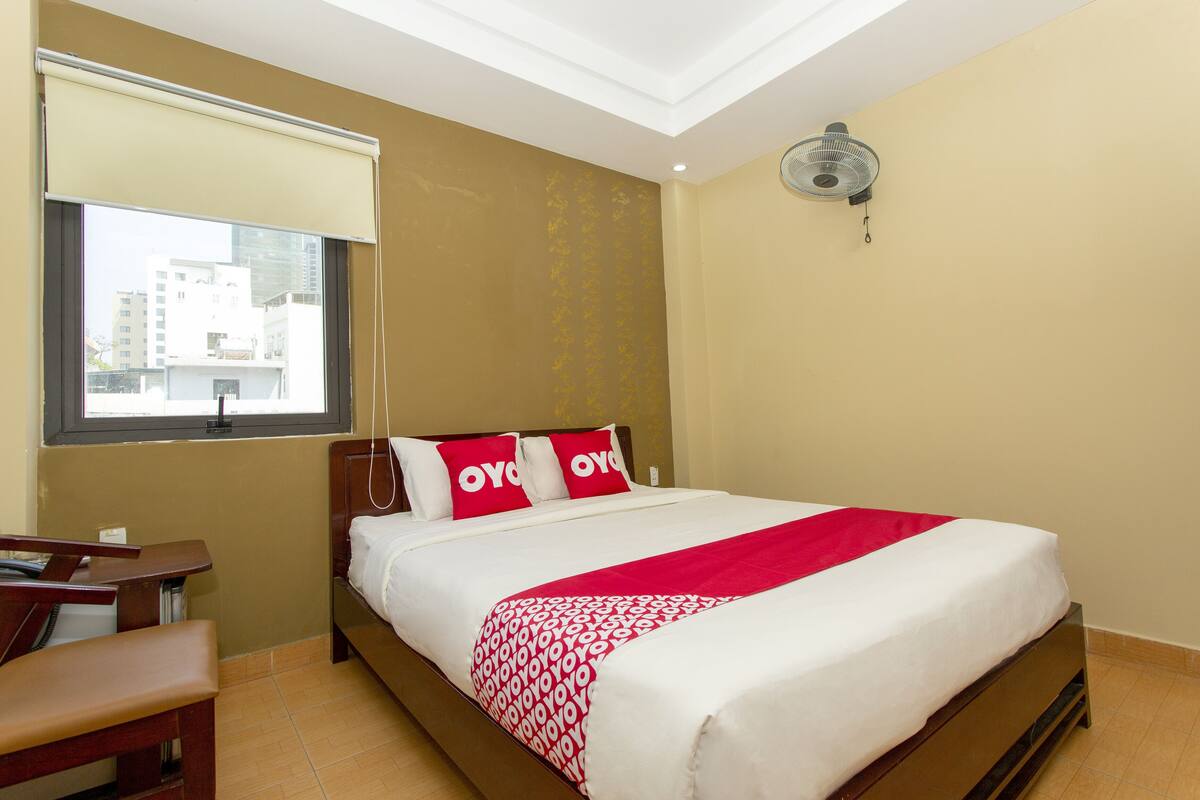 Een hotelkamer met een bed, een bureau, een stoel, een raam met uitzicht op gebouwen, en een plafondventilator.
