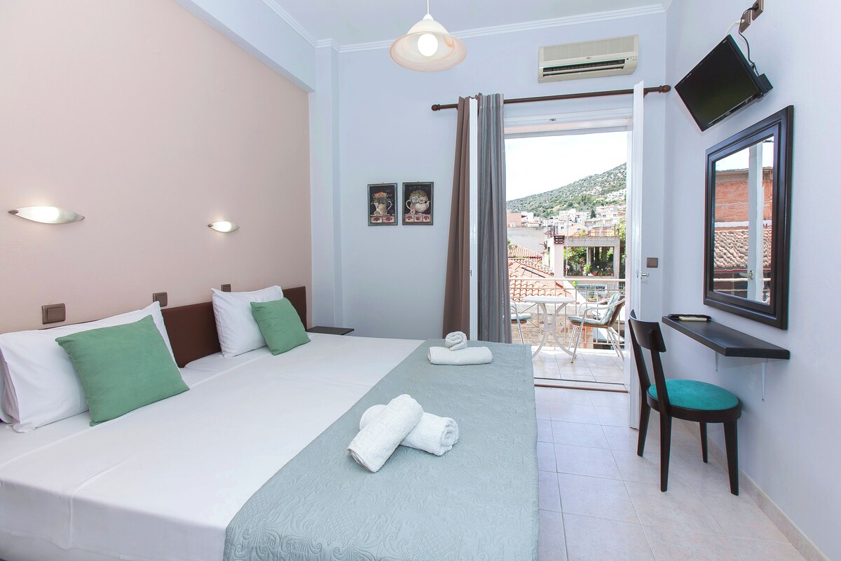 A hotel room with a bed, a desk, a chair, a TV, and a balcony with a view of buildings and hills.