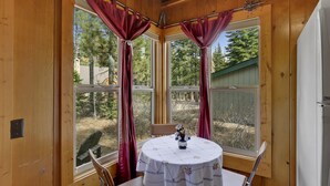Rincón acogedor con una mesa y sillas, ventanas grandes con cortinas y vistas a árboles y un edificio.
