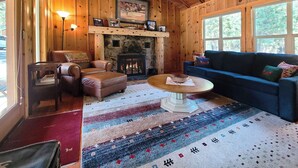 Un interior de cabaña acogedor con chimenea de piedra, mobiliario de cuero y ventanas amplias.