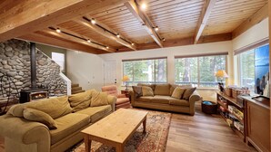 A cozy living room with a stone fireplace, wooden ceiling beams, and large windows offering a view of the outdoors. - Clubhouse - 4 bedroom, 3 bath - hot tub, quiet location (Tahoe City)