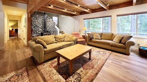 A spacious living room with a stone fireplace, wooden ceiling beams, and large windows offering a view of the forest. - Clubhouse - 4 bedroom, 3 bath - hot tub, quiet location (Tahoe City)