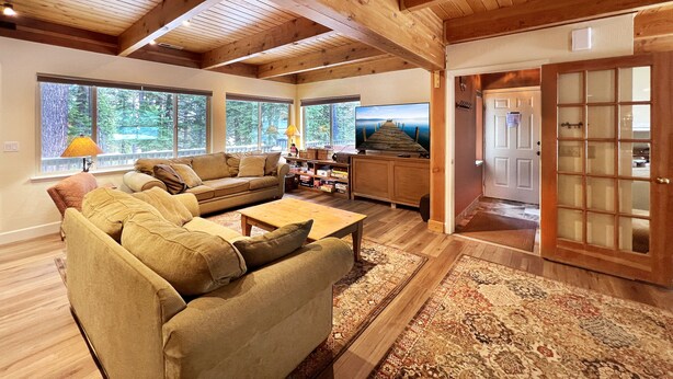 A spacious living room with a large sofa, a coffee table, and a television. There are large windows providing a view of the outdoors. - Clubhouse - 4 bedroom, 3 bath - hot tub, quiet location (Tahoe City)