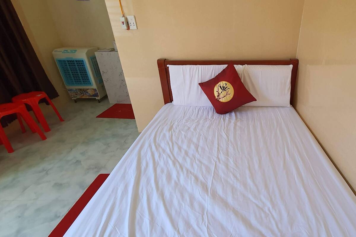 A room with a bed, a red stool, and an air cooler.