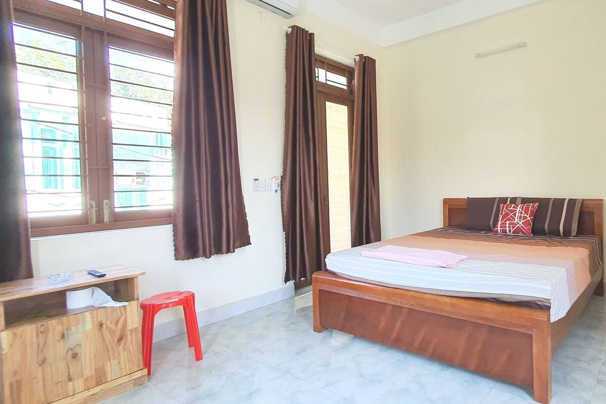 A bedroom with a wooden bed, a small wooden desk, a red stool, and a window with curtains.
