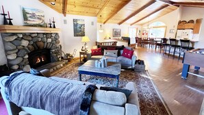 A cozy living room with a stone fireplace, wooden ceiling beams, and a dining area with a bar. - Chalet Lani - HOT TUB - 3 bedroom, 2 bath - Quiet Location! (Homewood)