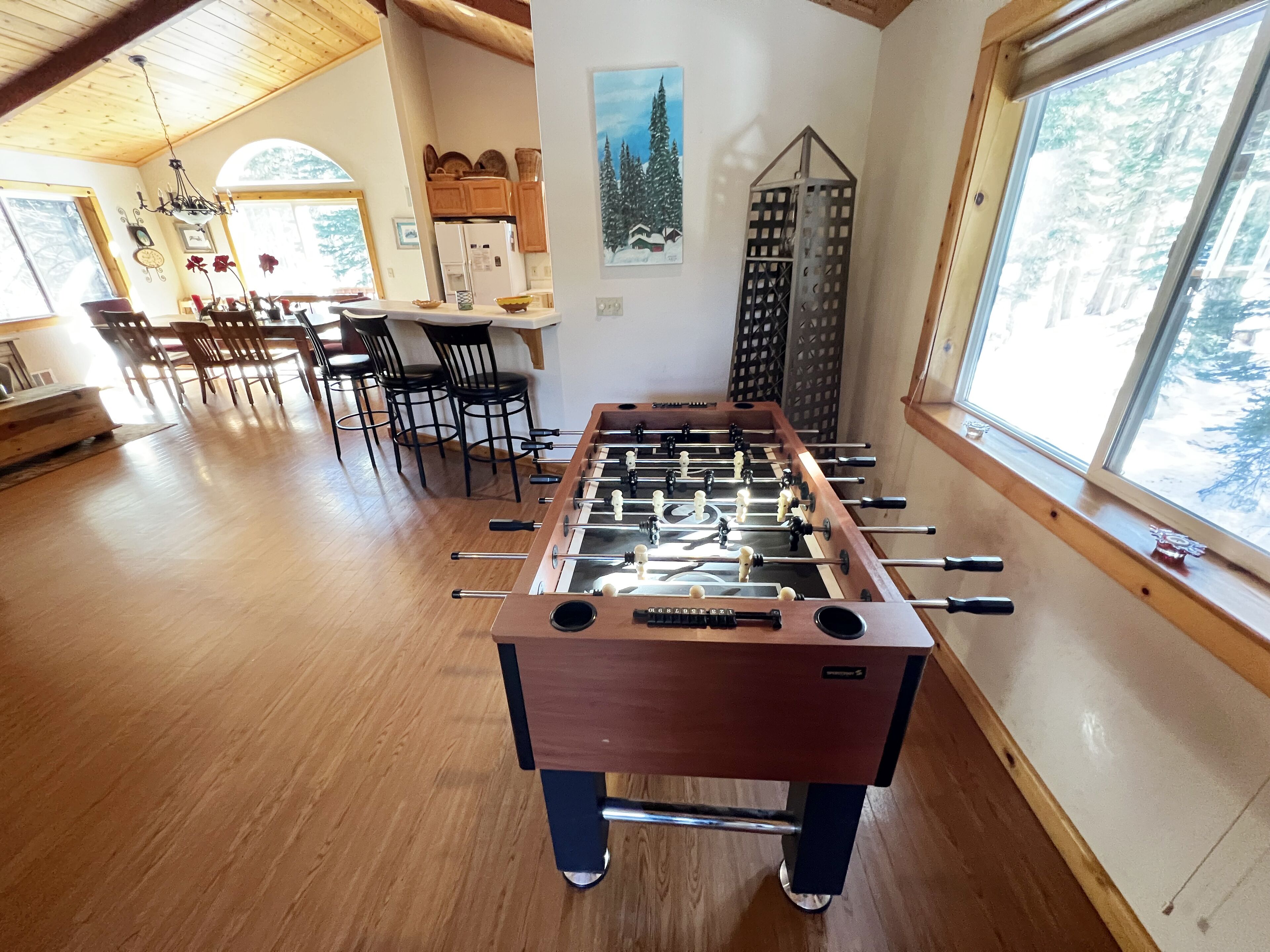 Ein Zimmer mit Tischfußball, Holzboden, einer Essecke mit Stühlen und Tisch sowie einem Fenster mit Blick auf Bäume.