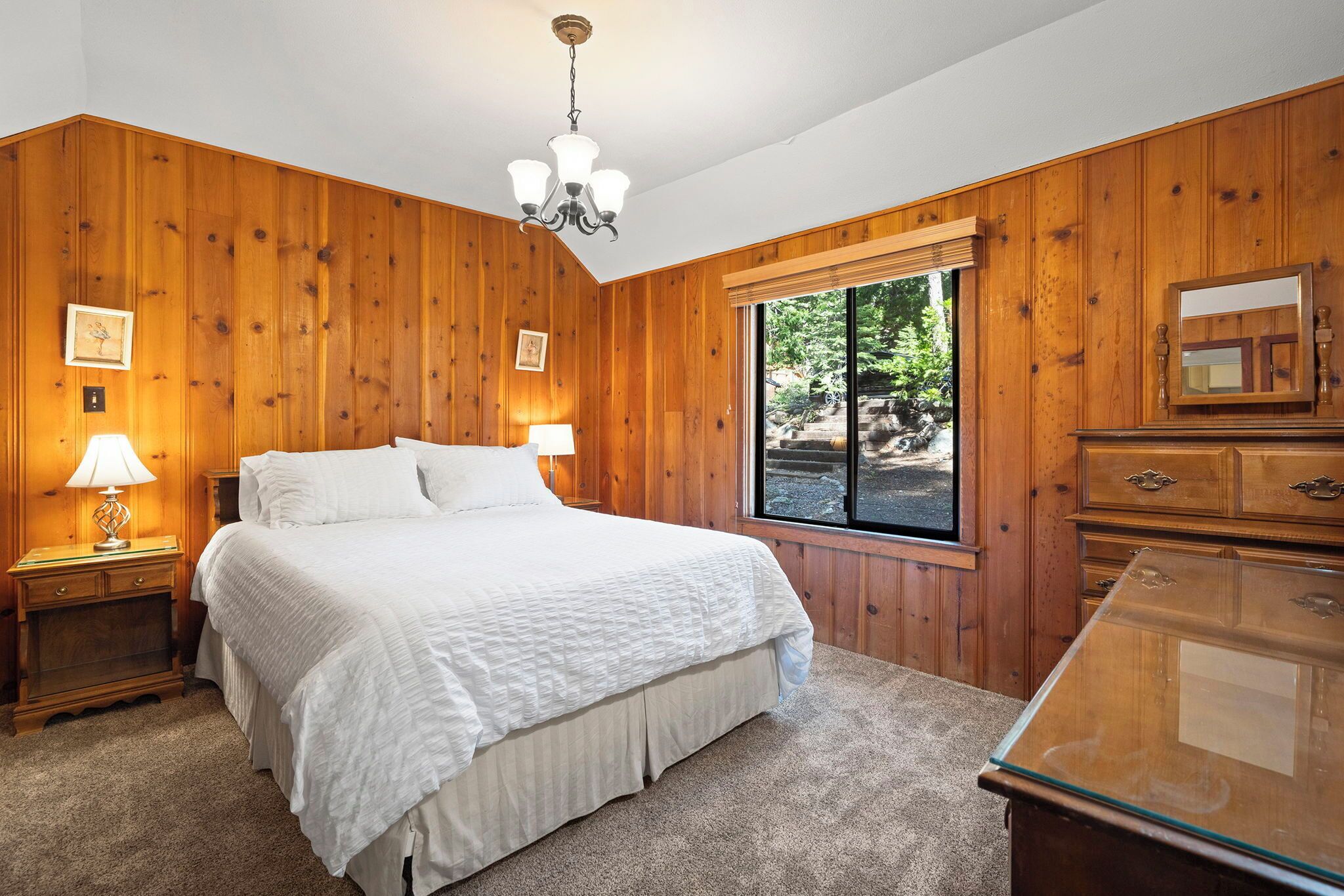 Een slaapkamer met een houten wandpaneel, een groot bed met wit beddengoed, een nachtkastje met een lamp, een dressoir en een raam met uitzicht op bomen.