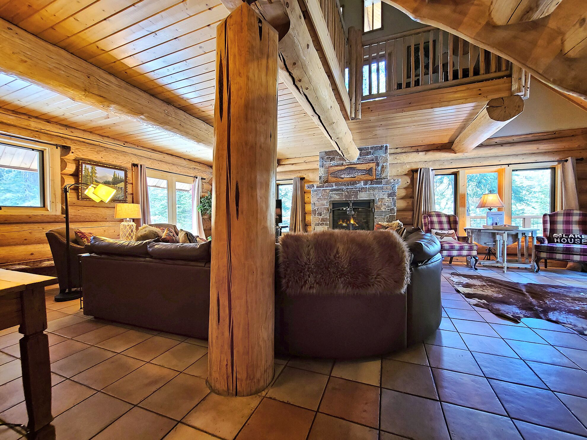 A cozy log cabin interior with a stone fireplace, wooden furniture, and large windows.