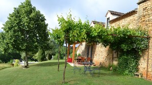 Un edificio de piedra con una pérgola cubierta de parras, un césped y una zona de comedor con mobiliario exterior.