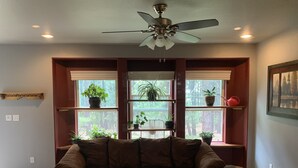 A living room with a brown leather couch, a ceiling fan, and shelves with potted plants. - Peace and privacy await among the towering Pine Trees. (Bayfield)