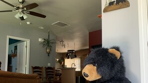 A living room with a large teddy bear holding a 'WELCOME' sign, a ceiling fan, a kitchen in the background, and a 'WELCOME' sign on the wall. - Peace and privacy await among the towering Pine Trees. (Bayfield)