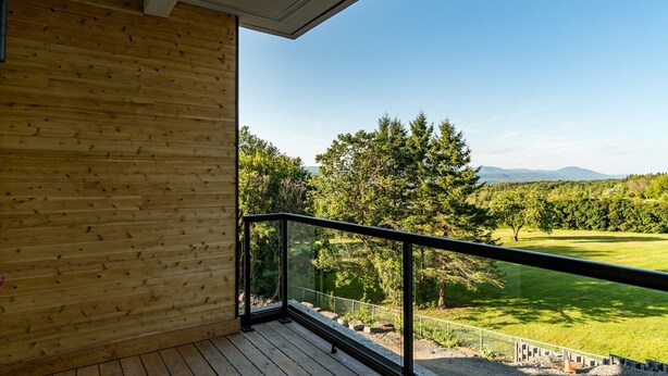 Un balcon en bois doté d’une rambarde en verre, qui offre une vue sur un paysage verdoyant, avec des arbres et des montagnes.