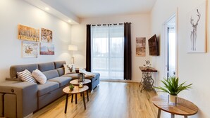 Een moderne woonkamer met een grijze bank, een salontafel en een bijzettafeltje. Er hangen kunstwerken aan de muur en er staat een televisie.