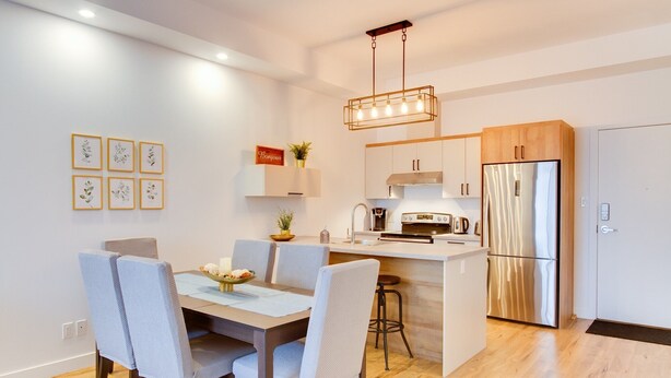 A modern kitchen with white cabinets, a stainless steel refrigerator, and a dining area with a table and chairs. - L'île-Do | condos neufs sur l'ile d'Orléans, The Bouquet - 801 (Saint-Pierre-de-l'Île-d'Orléans)