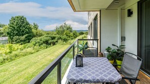 A balcony with a table and chairs, potted plants, and a view of a green landscape. - L'île-Do | condos neufs sur l'ile d'Orléans, The Bouquet - 801 (Saint-Pierre-de-l'Île-d'Orléans)