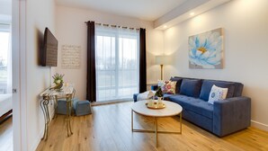A modern living room with a blue sofa, a round coffee table, and a large window with curtains. - L'île-Do | condos neufs sur l'ile d'Orléans, The Bouquet - 801 (Saint-Pierre-de-l'Île-d'Orléans)