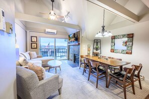 A modern living room with a sofa, a dining table, and chairs. There is a fireplace and a large window with a view.