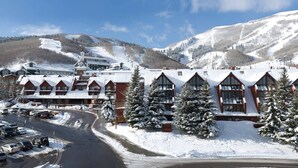Un chalet enneigé composé de plusieurs bâtiments, avec une piste de ski en arrière-plan et un parking où sont garées des voitures.