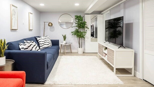 Una sala moderna con un sofá azul oscuro, una alfombra blanca, un sillón rojo, un televisor sobre un soporte blanco y un espejo grande en la pared.