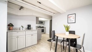 Una cocina moderna con armarios blancos, microondas y cafetera. Hay un comedor con una mesa redonda y cuatro sillas. Una planta en maceta está sobre la mesa.