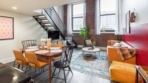 A modern living room with a dining area, a staircase, and large windows. - Sosuite | 3BR Loft w Gym, Lounge, Shared Laundry (Philadelphia)