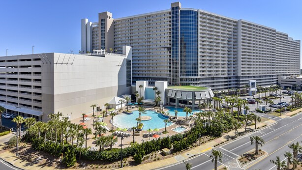 A large hotel with a central swimming pool, multiple parking levels, and a well-maintained landscaped area with palm trees.