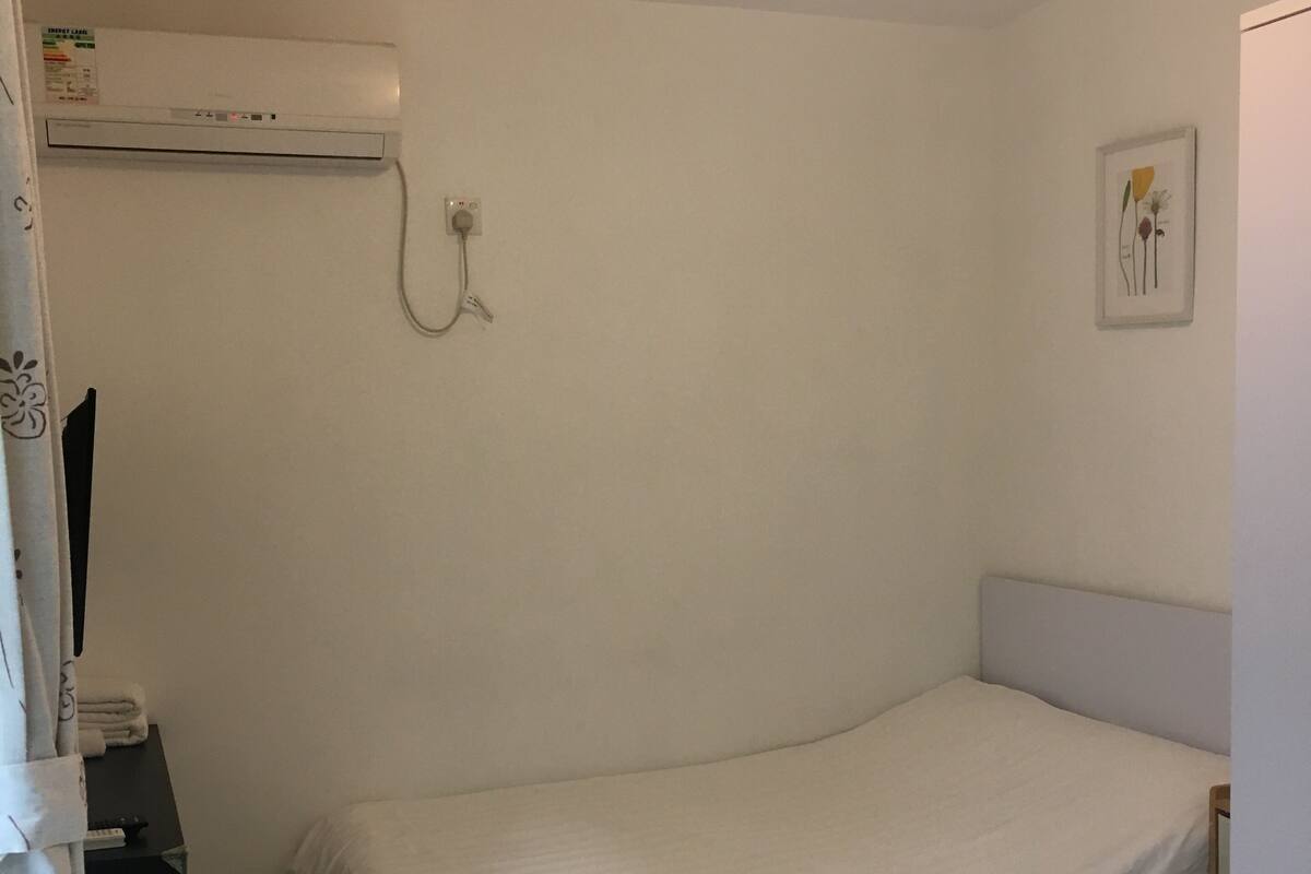 Un dormitorio pequeño y ordenado con una cama, una mesita de noche con una tetera y taza, una pequeña mesa con una taza y un aire acondicionado instalado en la pared.