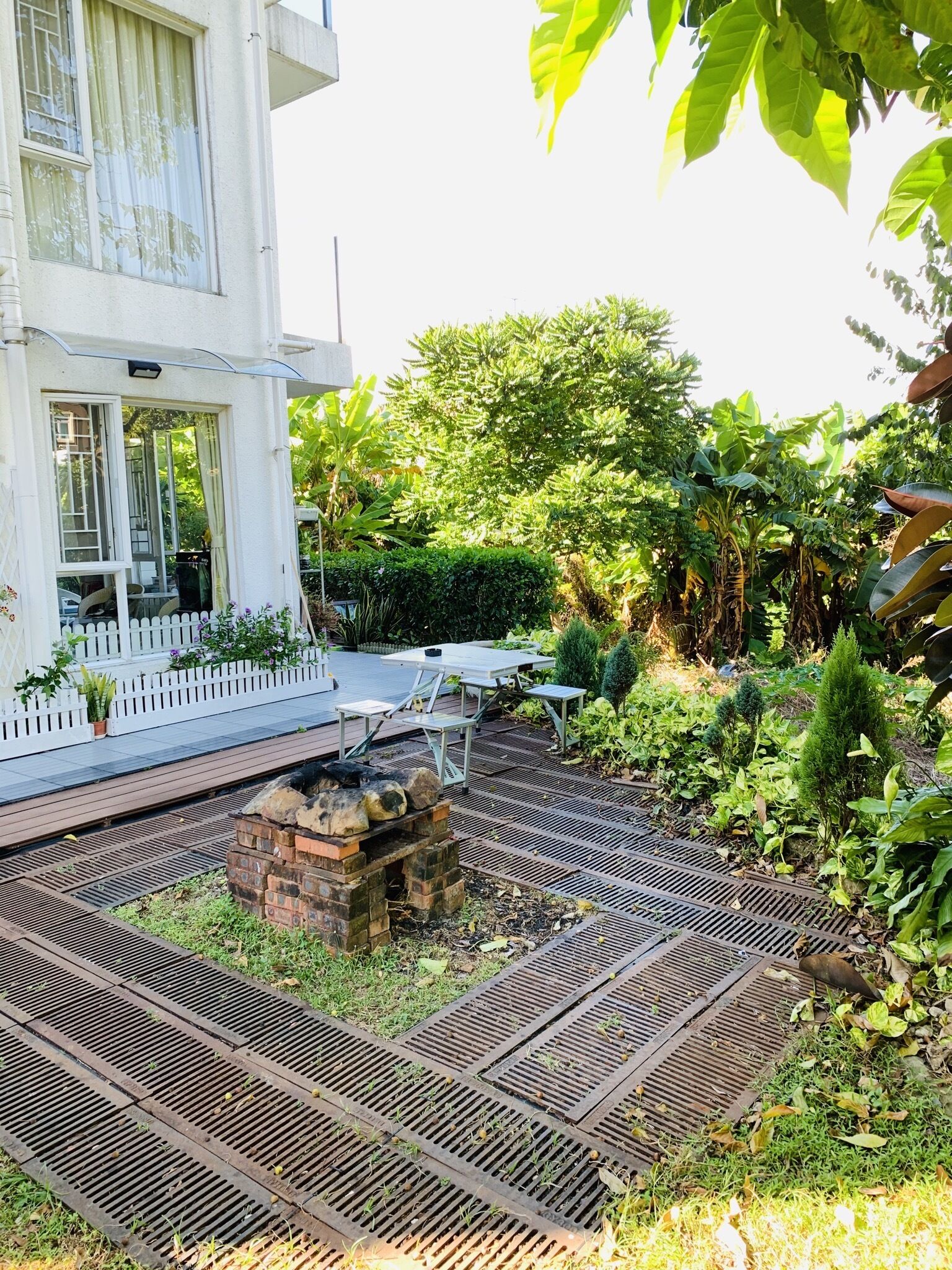 Een tuin met een vuurkorf van baksteen, picknicktafels en een witte omheining.