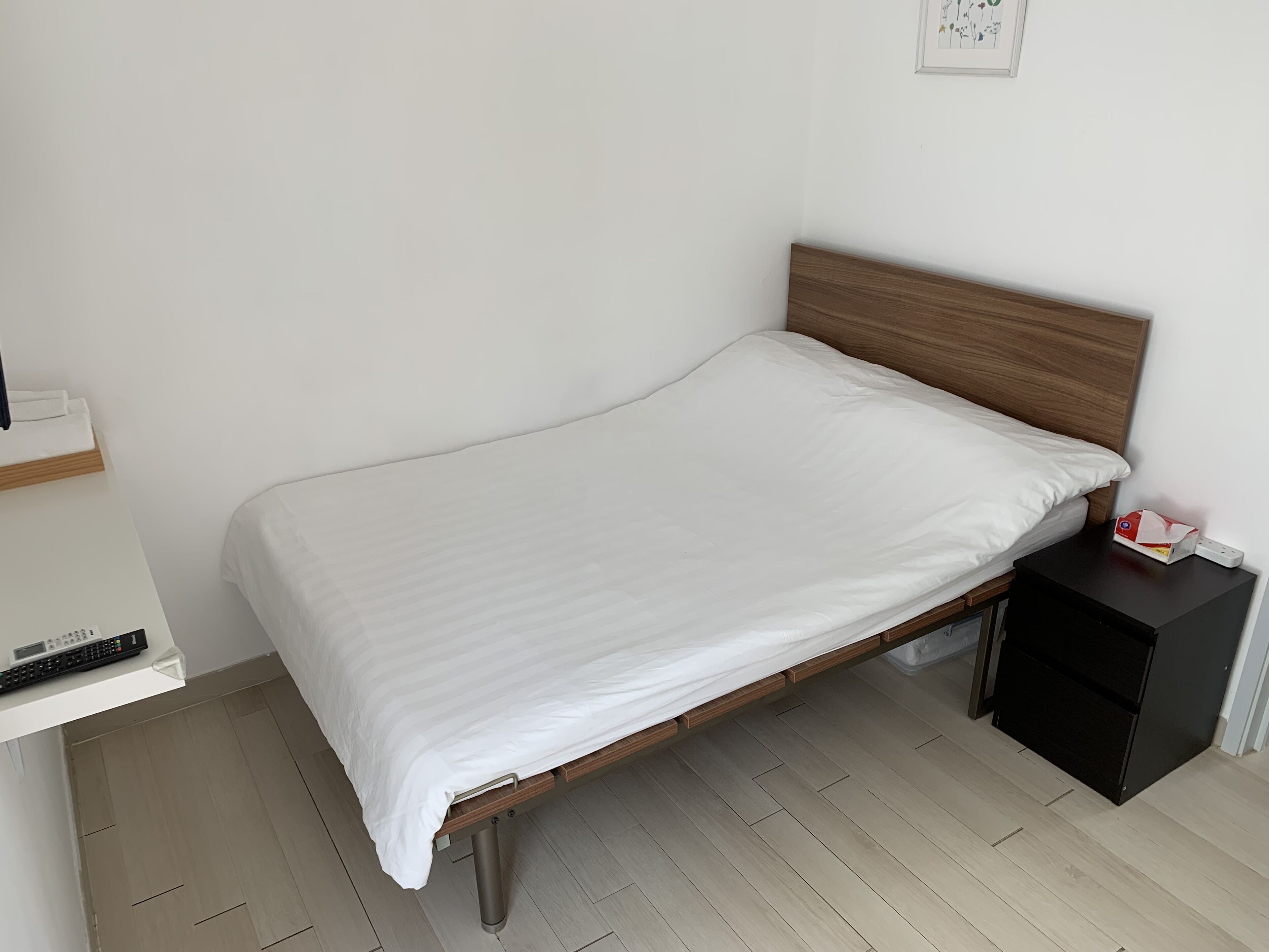A single bed with a wooden headboard, a bedside table with a tissue box, and a small shelf with a remote control.