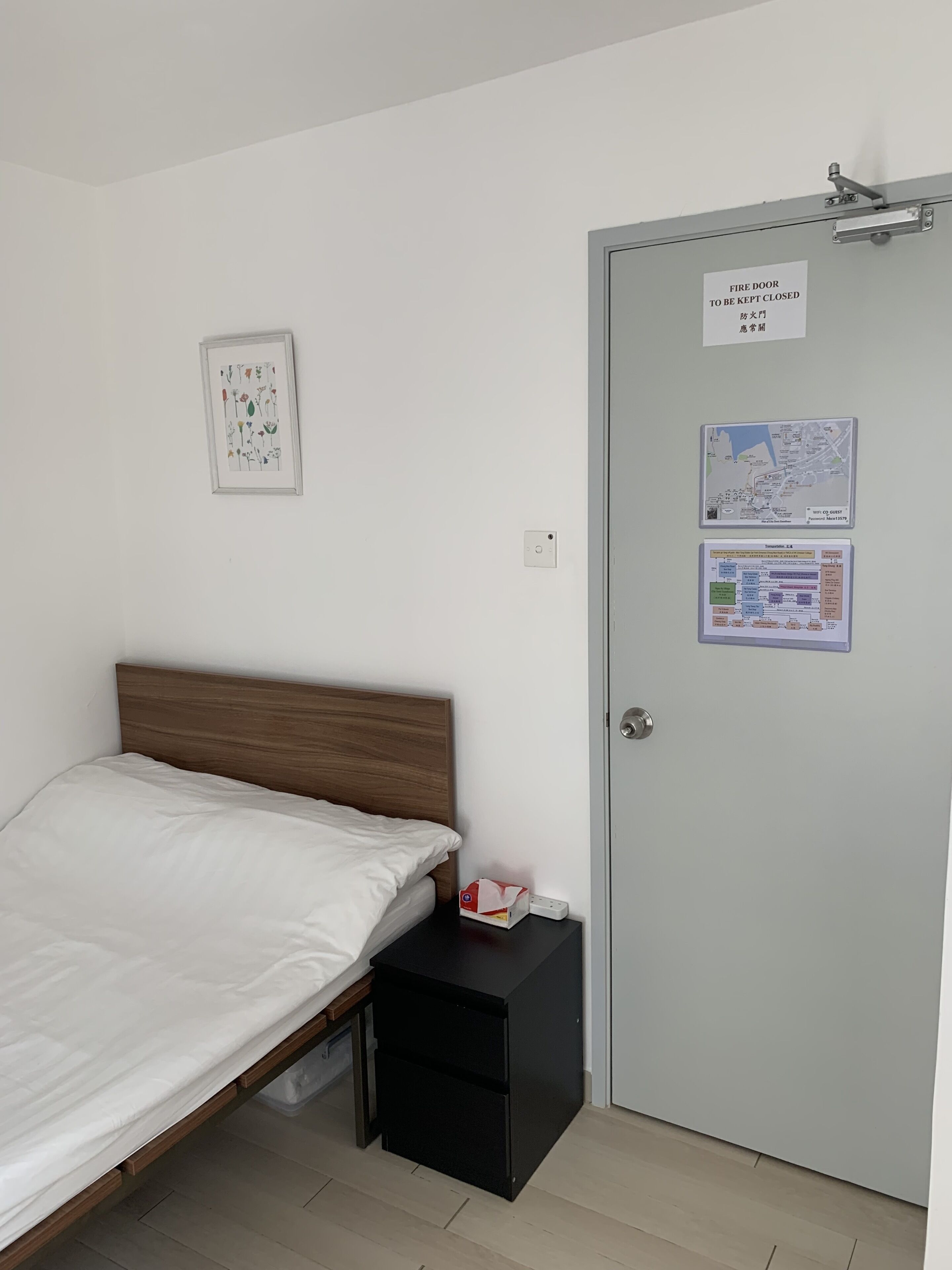 A bedroom with a bed, a nightstand, a door with a fire safety sign, a framed botanical print, and a map on the wall.