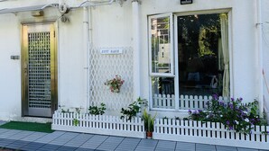 Un bâtiment à l’extérieur blanc, une porte en métal avec un motif en forme de grille, une enseigne indiquant « CITY OASIS », et une clôture blanche en bois avec des plantes en pot.