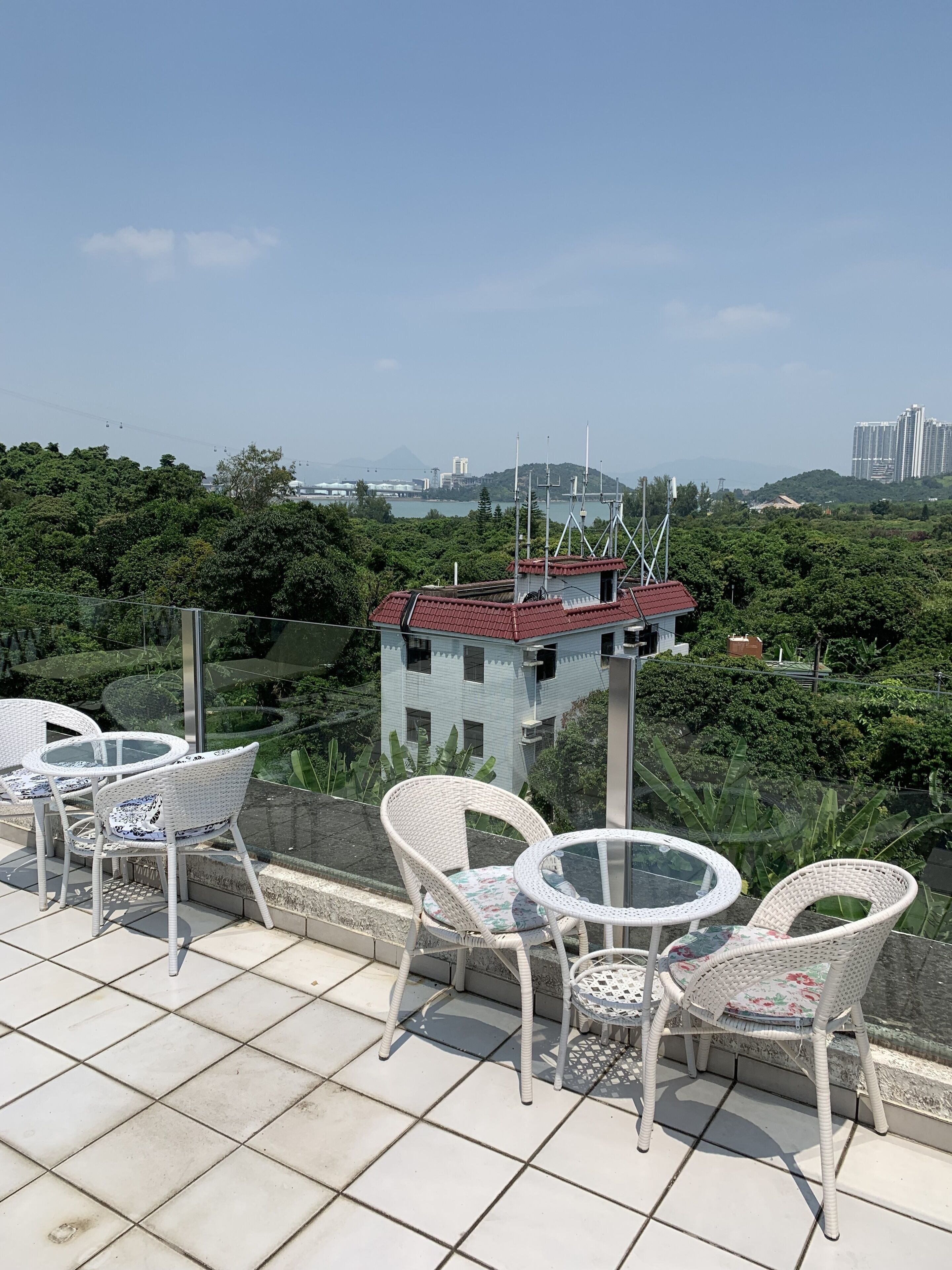 A terrace with white wicker chairs and a glass table, overlooking a cityscape with buildings and greenery.