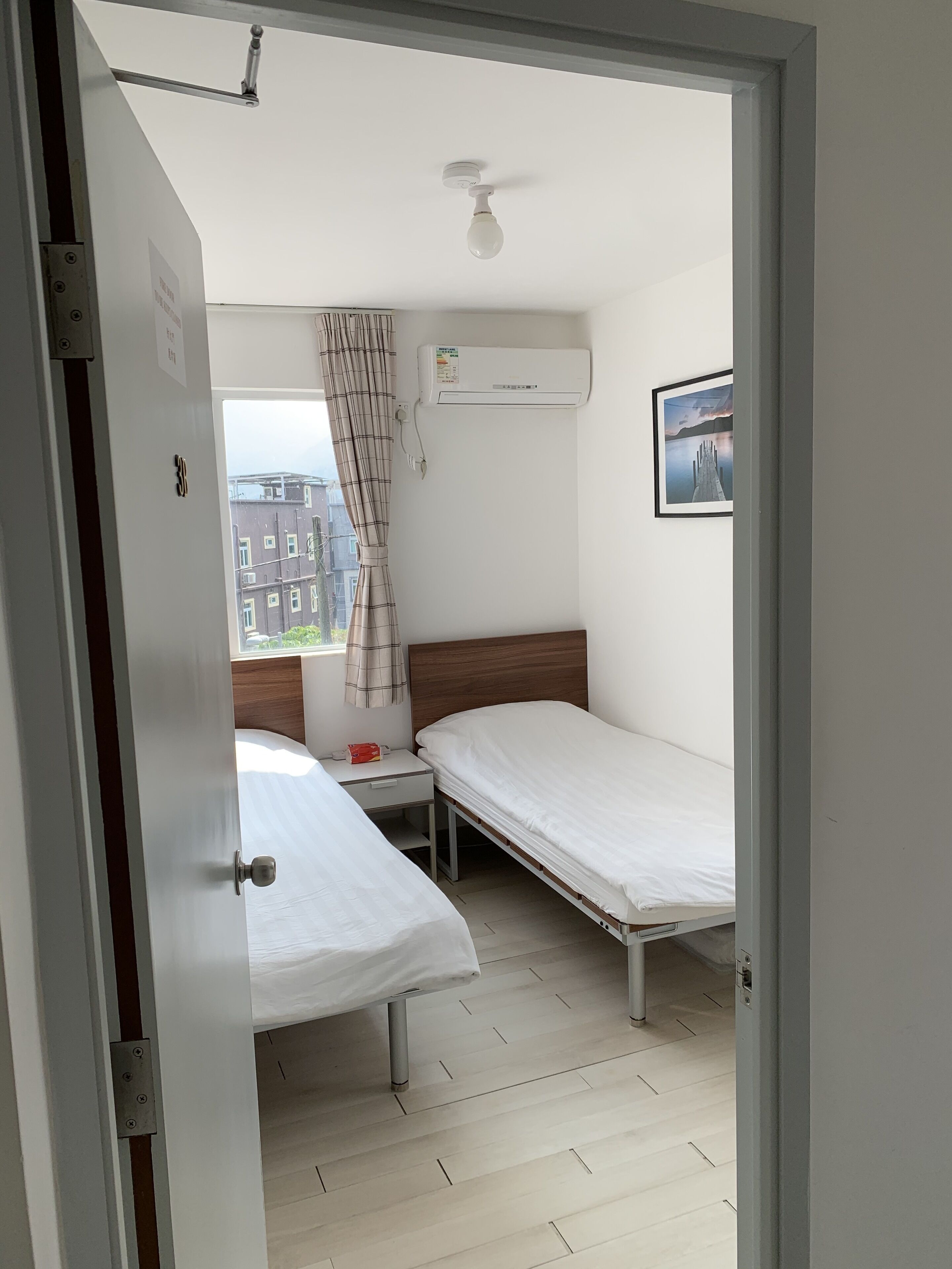 A bedroom with two single beds, a small bedside table, a wall-mounted air conditioner, and a window with a view of buildings.